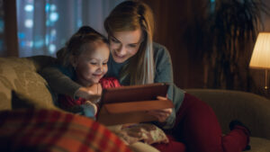 Beautiful Mother and Her Little Daughter are Sitting on a Sofa in the Living Room, They Use Tablet Computer. It's Evening, Room is Cozy and Warm.