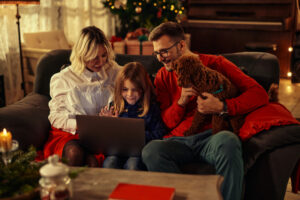 Little boy sitting between his parents on sofa in the living room and using laptop. Father holding poodle in his lap